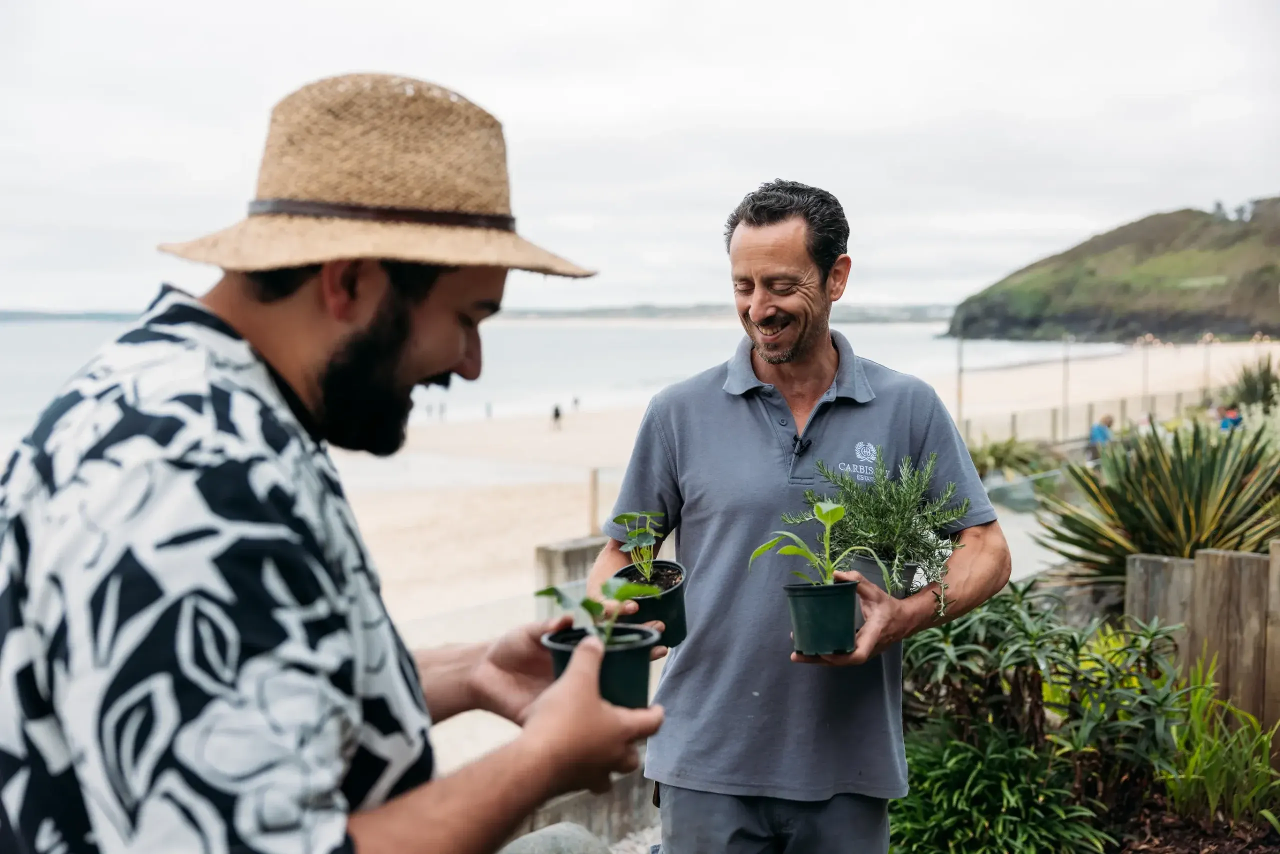 Chris the head gardener being interviewed by Joe from Joe's Garden TikTok channel at Carbis Bay Estate, holding fresh herb plants with the beach and Cornish coastline behind them