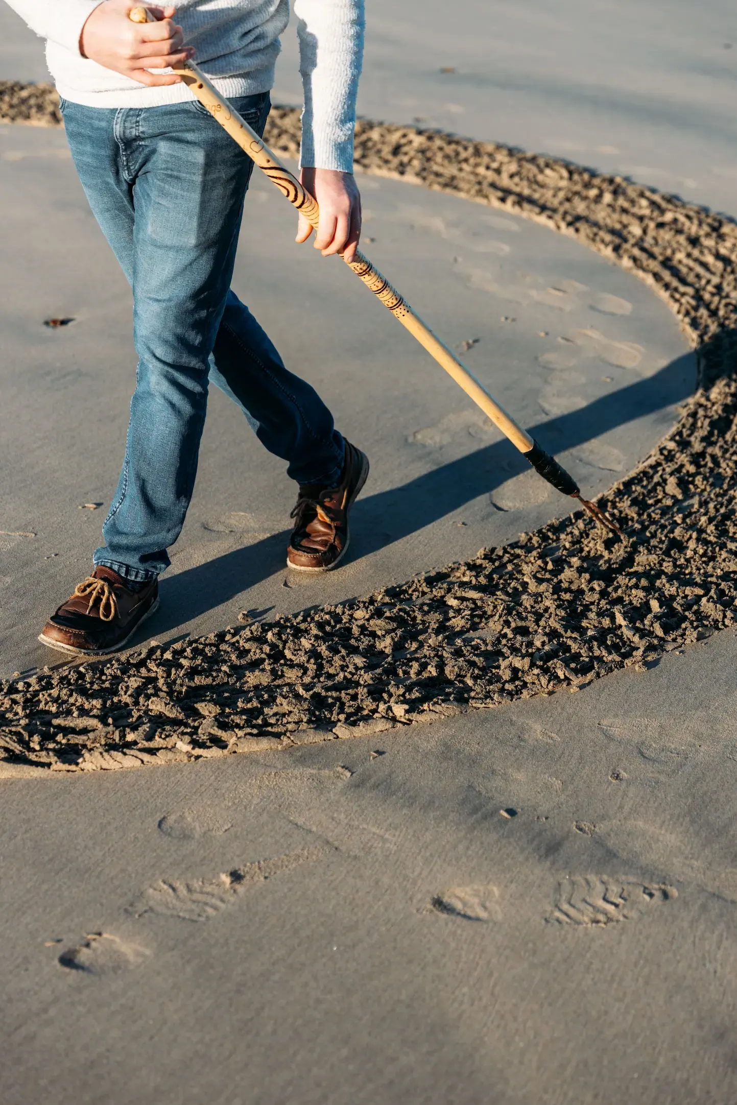 Sand artist drawing a large circular shape in the sand on a Cornish beach, using a wooden rake, photographed by PixelRain