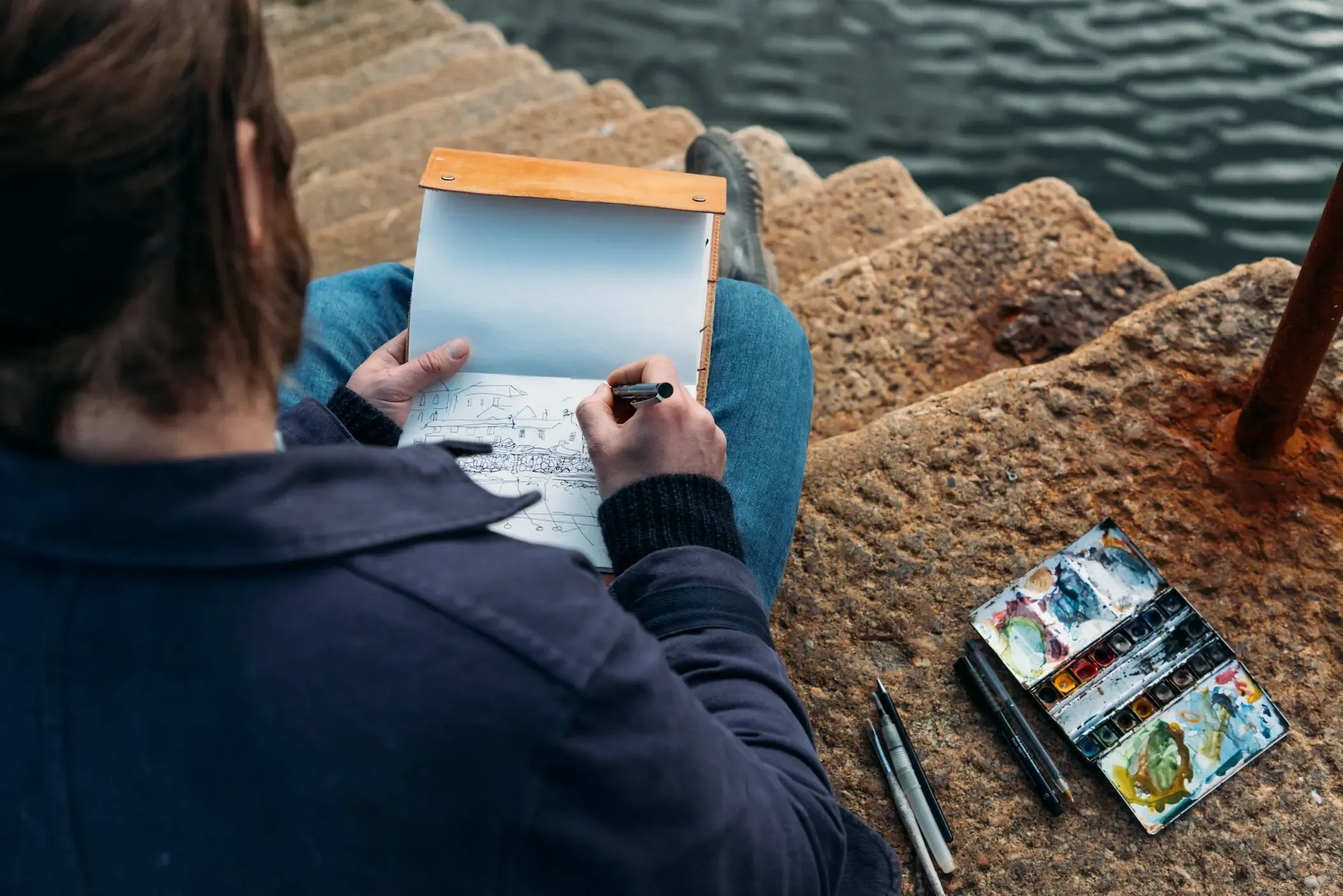 James Strawbridge seated on the harbour steps at Mousehole, sketching the village with a watercolour palette beside him, during production of Forgotten Cornwall for History Hit