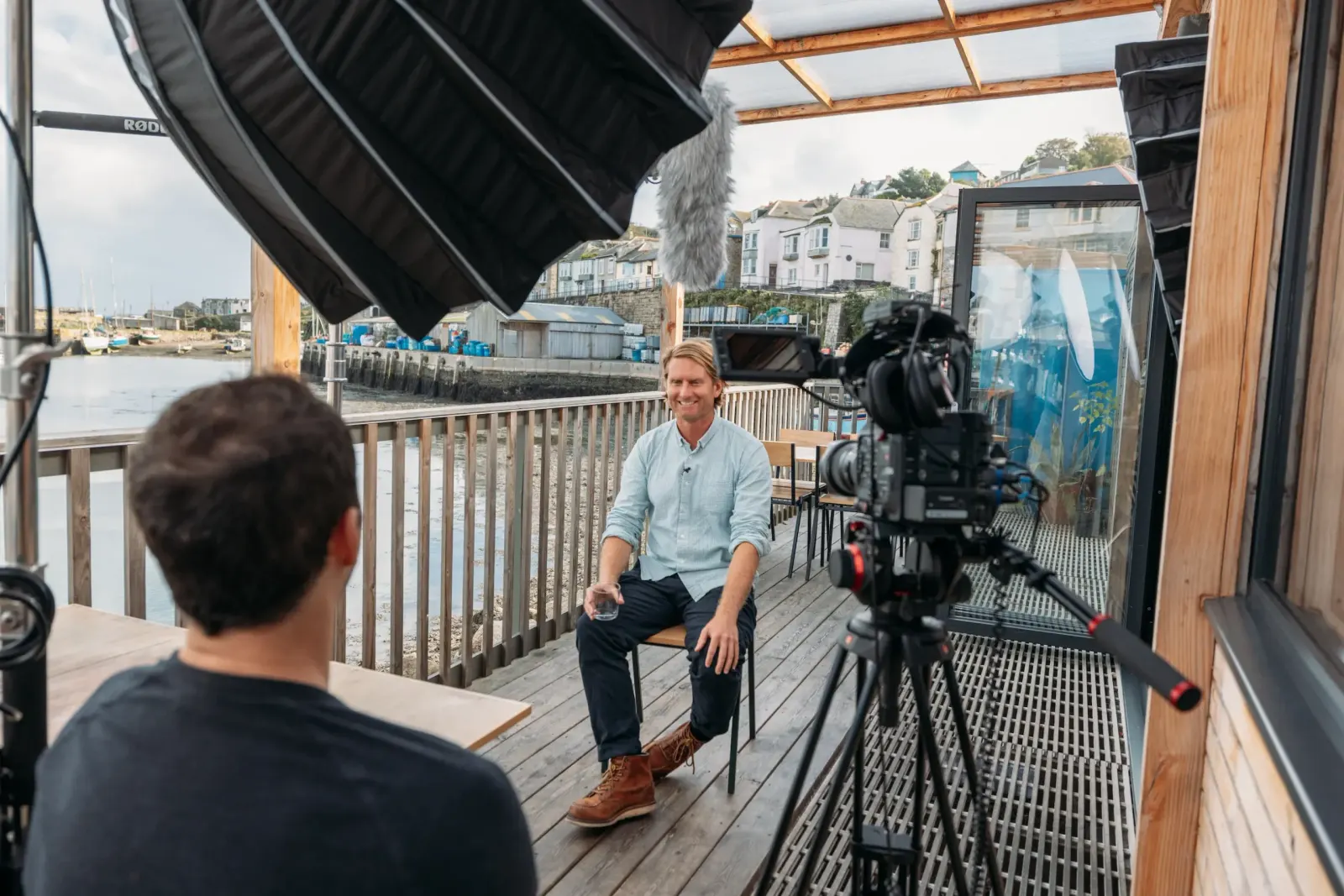 Behind the scenes of a PixelRain on-camera interview at Newlyn harbour, Cornwall, showing the interviewee seated on a waterfront deck with cinema camera, boom microphone, and professional lighting setup