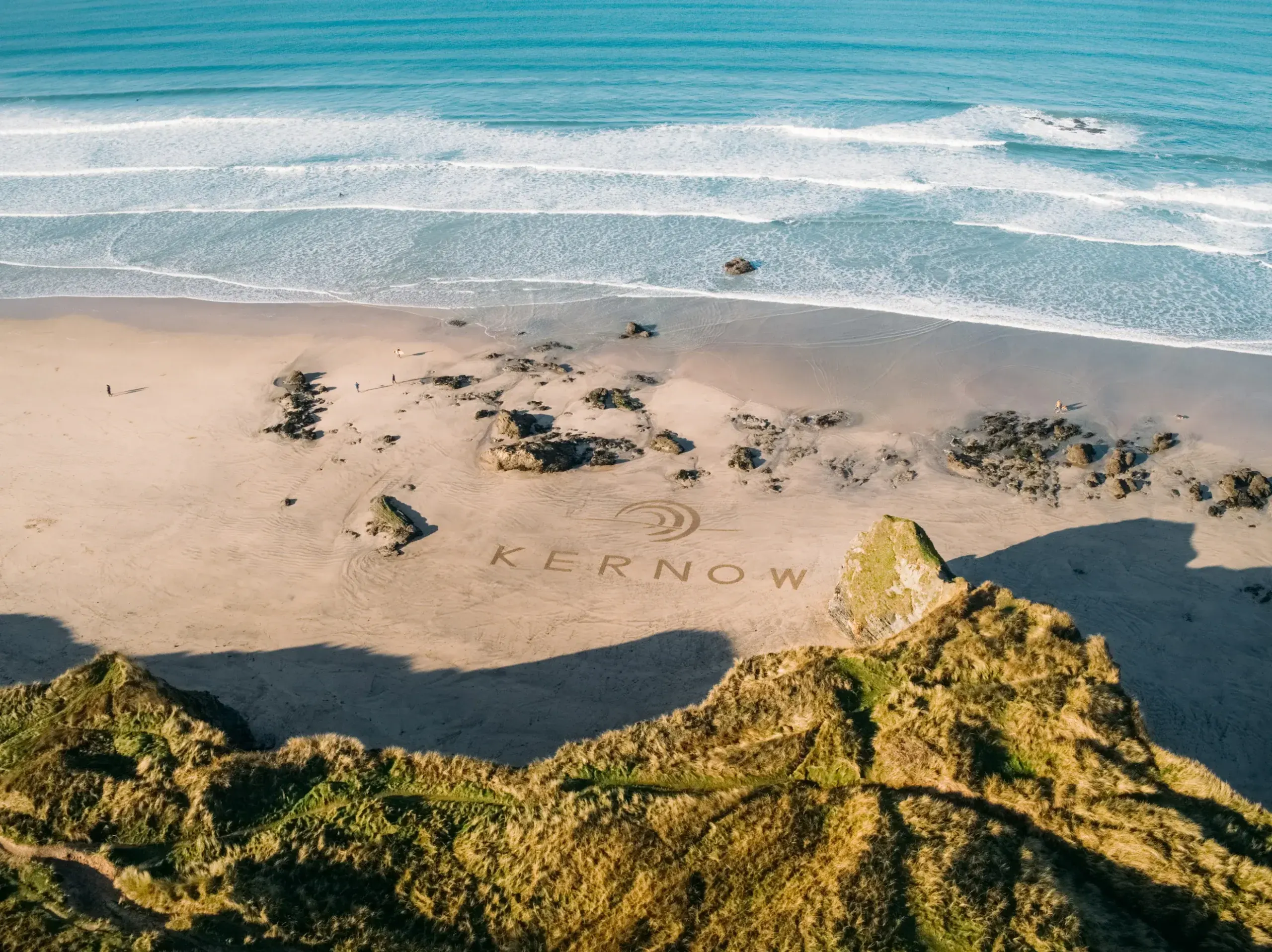 Aerial view of the Kernow Asset Management logo drawn in sand on a Cornish beach, with Atlantic waves and coastal dunes, filmed by PixelRain